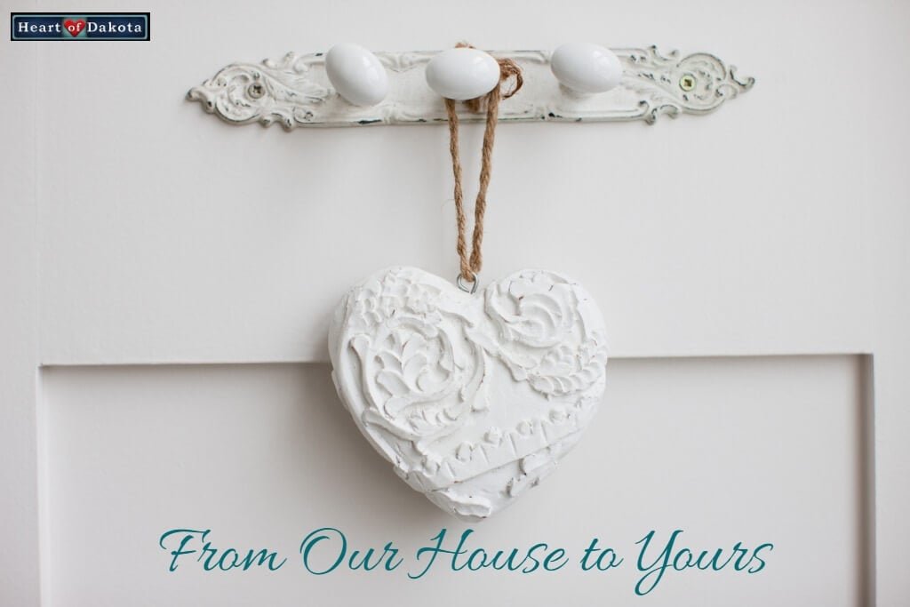 White painted, decorative plaster heart hanging in front of a white door. Blue text near the bottom reads: "From Our House to Yours."