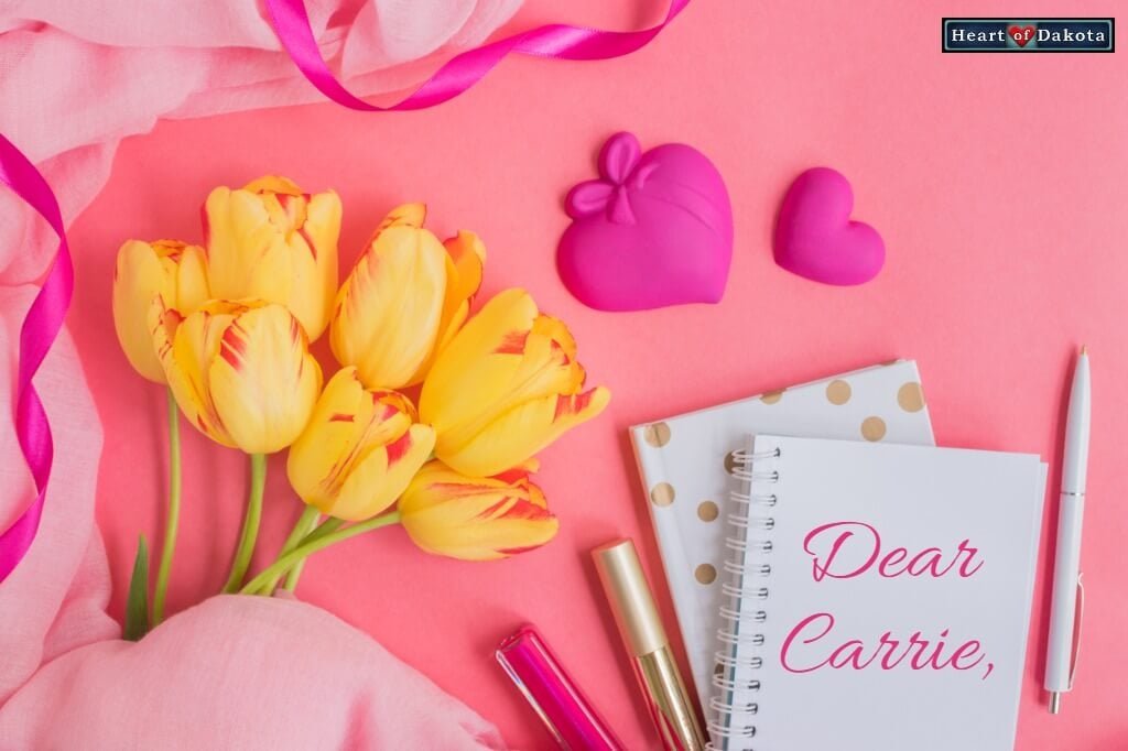 Small white notebook lying on a pink background besides yellow tulips. On the notebook, pink script reads "Dear Carrie."