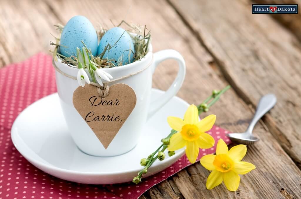 White ceramic cup with robin eggs and green grass inside. A cardboard paper heart is tied to the cup with twine and has "Dear Carrie" written on it.